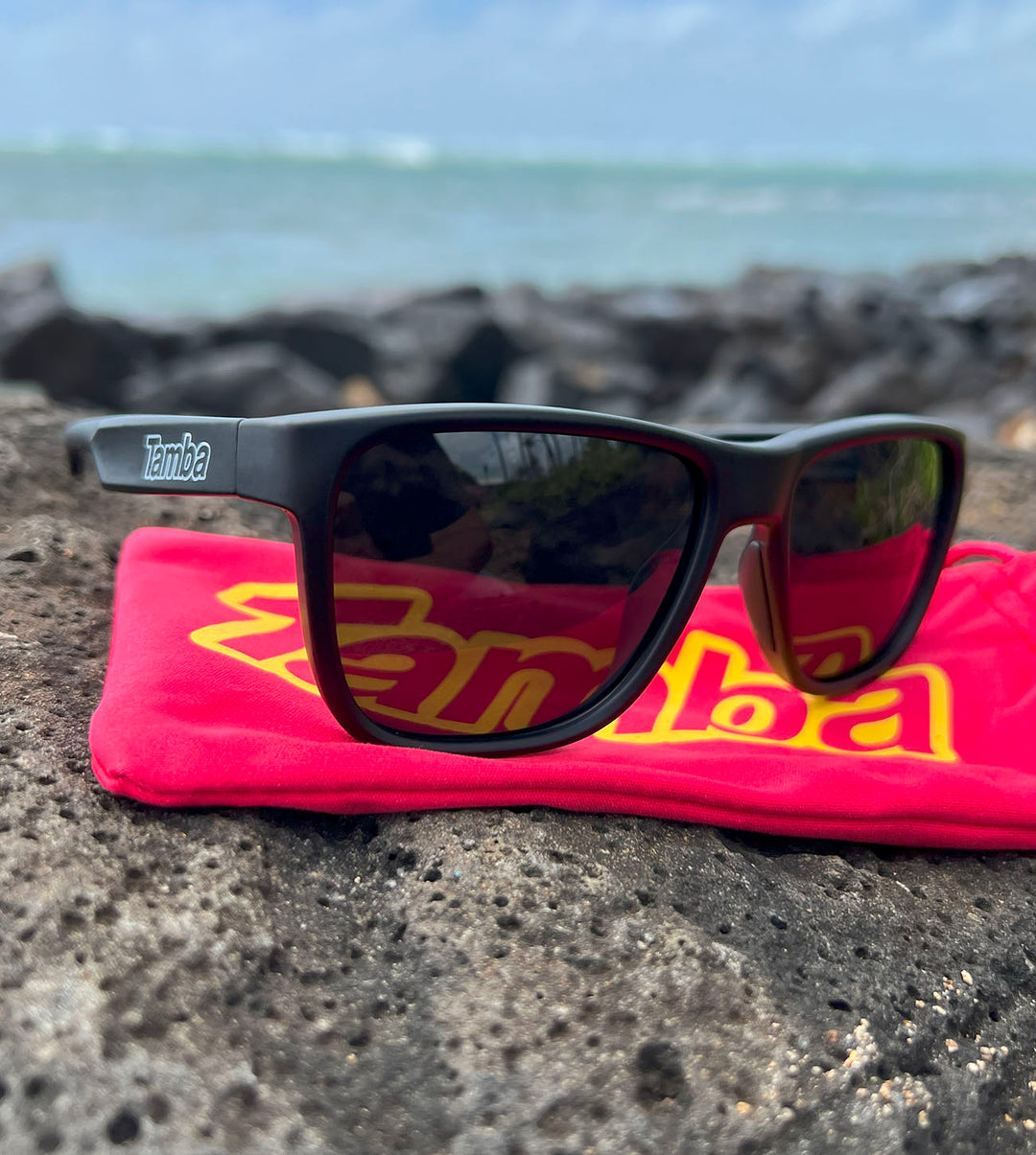Tamba Sunglasses (Matte Black) sitting on a rock by a beach.
