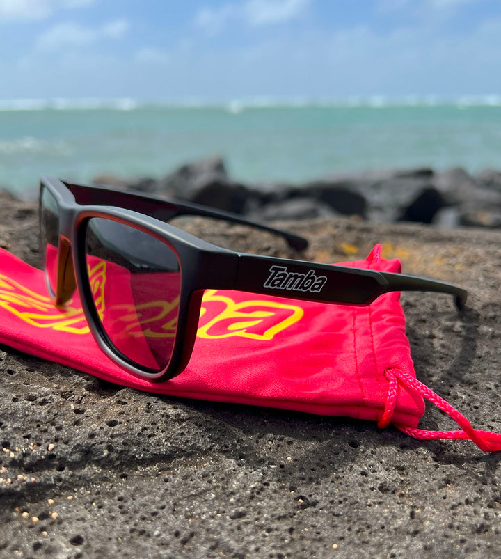 Tamba Sunglasses (Matte Black) sitting on a rock by the beach with the arm logo showing.